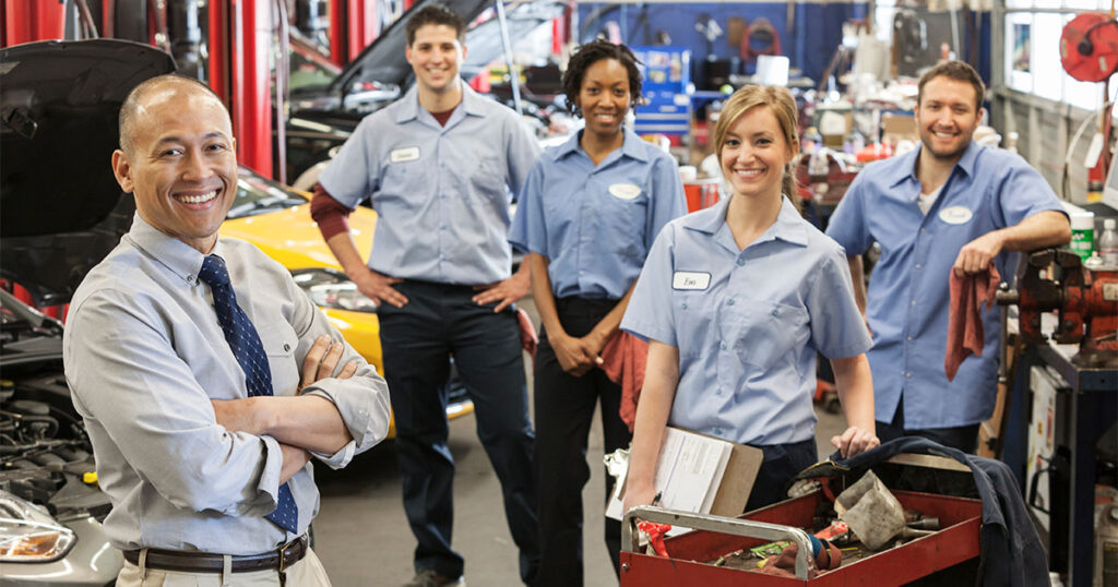 A group of auto mechanics are smiling and posing inside of an automotive repair shop.